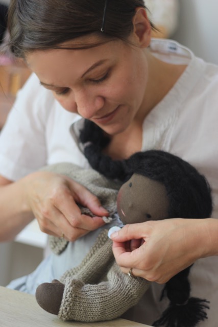 Über die Autorin: Maike knöpft einer Puppe mit schokobrauner Haut die beige Strickjacke zu
